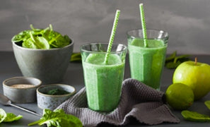 This is a picture of two green smoothies in glass cups with a green apple and a bowl of greens on a grey table with a grey background.