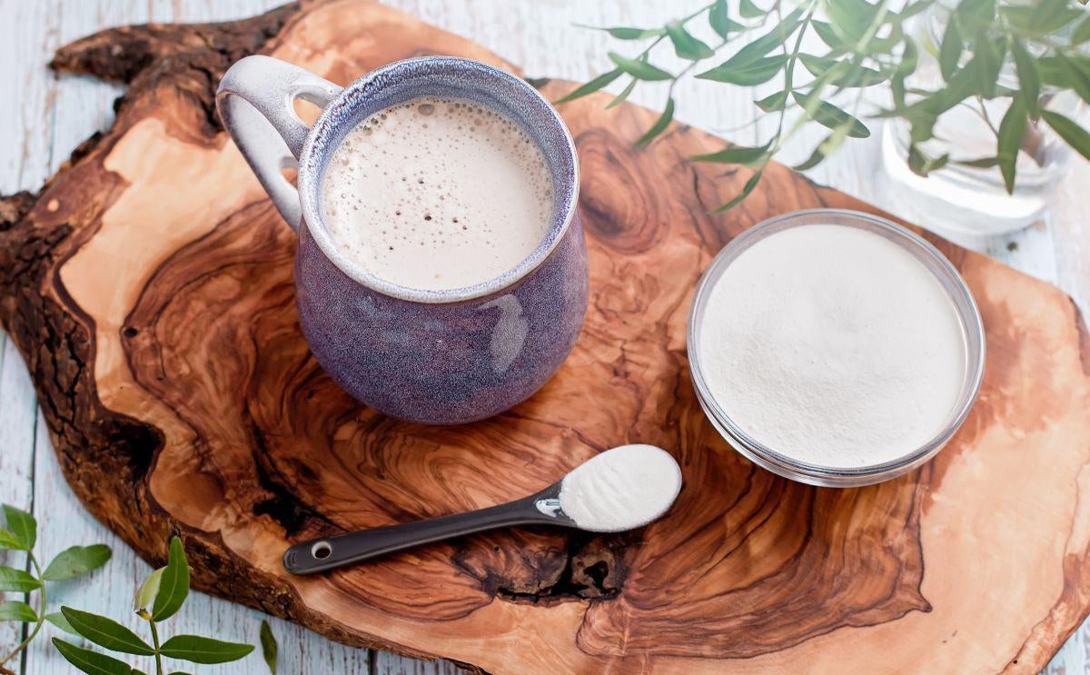 This is a picture of a cup of coffee in a blue mug, next to a glass bowl of collagen powder and a serving spoon sitting on a wooden serving board, on top of a wooden table top.