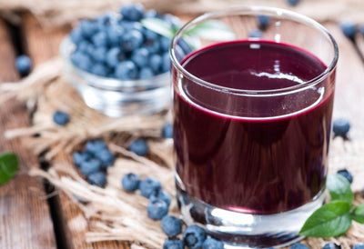 This is a picture of a glass of blueberry juice sitting next to a bowl of blueberries on a wooden table.