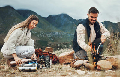 This is a picture of a happy man and woman cooking outside in a camping environment with the mountains in the background.