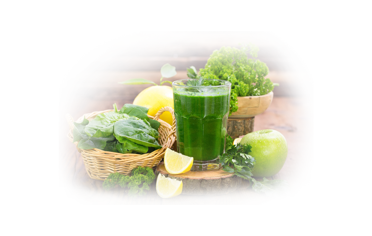 Image of clear glass containing green smoothie next to small basket filled with spinach leaves with a bowl of kale in the background.